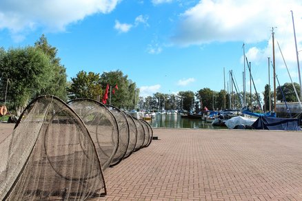 zeigt eine aufgezogene Reuse auf einer Pflasterfläche, im Hintergrund der Seglerhafen mit Segelbooten