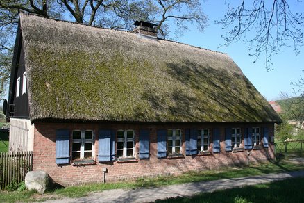 zeigt ein altes Bauernhaus aus Backstein, mit Schilfdach und blauen Fensterläden