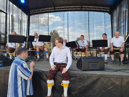 zeigt Bläsergruppe auf einer Bühne, davor Leif Tennemann und ein junger Mann in Lederhosen im Gespräch