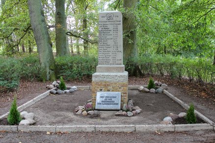 zeigt steinerne Säule und Tafel mit Inschrift sowie schlichte Flächeneinfassung aus Beton