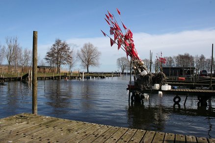 zeigt Blick auf eine Wasserfläche mit Bootsstegen aus Holz und einem Bund Reusenstangen mit rotem Wimpel, dahinter Schilf- und Grünfläche