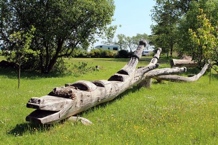 zeigt ein aus einem Baumstamm gestaltetes Krokodil auf einer Wiesenfläche