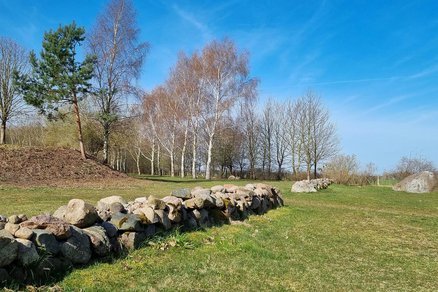zeigt aufgeschichtete Wälle aus Findlingsgestein auf einer hügeligen Grünfläche, im Hintergrund Baum- und Strauchbewuchs