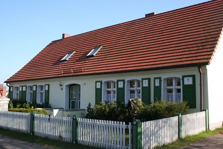 zeigt Wohngebäude mit hellem Putz, grünen Fensterläden und rotem Ziegeldach