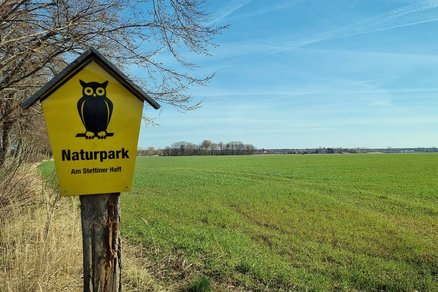 zeigt ein Landschaftspanorama, im Vordergrund ein Schild "Naturpark am Stettiner Haff" am Rande eines weiten grünen Feldes
