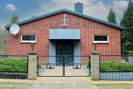 zeigt Frontseite mit Eingang einer kleinen Kirche aus Klinkermauerwerk, mit Zaun davor